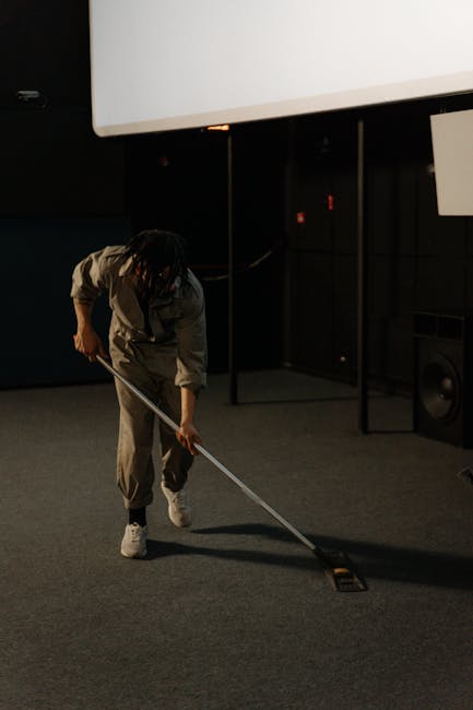 A person wearing grey overalls is engaged in surface cleaning in a darkened indoor environment, possibly a commercial space such as a cinema or auditorium. They are using a long-handled mop or cleanin